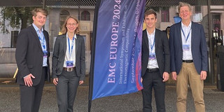 EMC_Europe2024_Bruegge_Gruppenfoto Drei wissenschaftliche Beschäftigte und Professor Frei sind auf dem Bild zu sehen. Sie stehen neben einer Fahne mit dem Schriftzug "EMC Europe 2024".